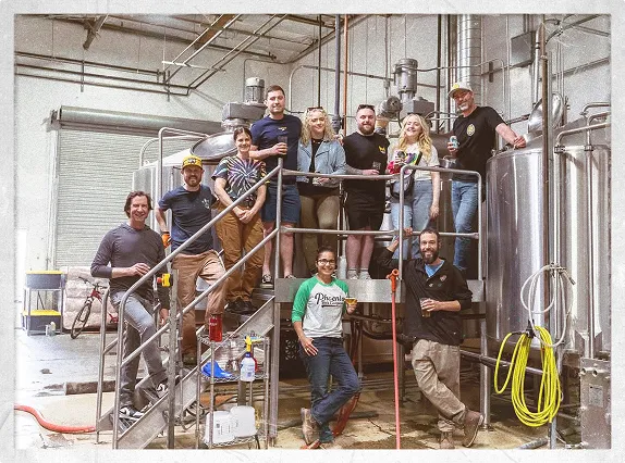 Mixed group of Phoenix Brewery and PHX Beer Co. staff gathered on and around a raised brewing platform inside a working brewhouse, smiling for a photo in front of large stainless-steel tanks and brewing equipment.