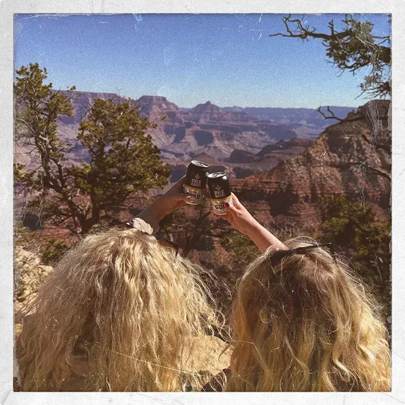 Two people with curly hair seen from behind raising PHX Beer Co. cans in a cheers gesture, overlooking the dramatic cliffs and rock formations of the Grand Canyon under a clear sky.