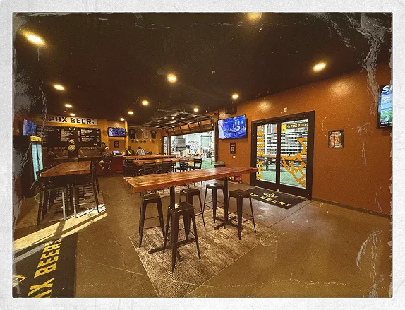 Warm, industrial-style taproom interior at PHX Beer Co. with high tables and bar stools, tiled bar counter, wall-mounted TVs and branded signage, with large glass doors leading through into the brewery area.
