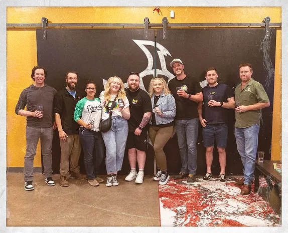 Group photo of brewery staff lined up indoors against a black wall with a phoenix logo, holding drinks and smiling after a visit to the taproom.