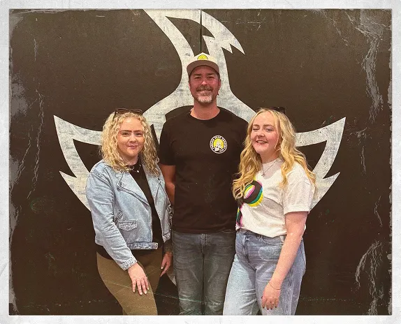 Three people standing together in front of a large black wall painted with a stylised phoenix emblem, smiling casually for the camera.