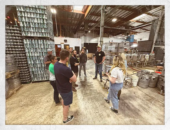Group of brewery staff standing in a circle inside a large production warehouse, surrounded by towering stacks of empty aluminium cans and metal kegs while listening to a team member speak.