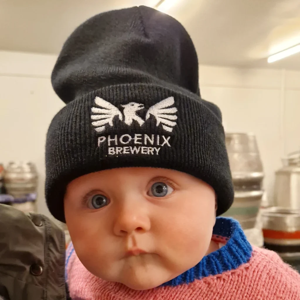 Baby wearing an oversized black Phoenix Brewery beanie hat with white embroidered logo