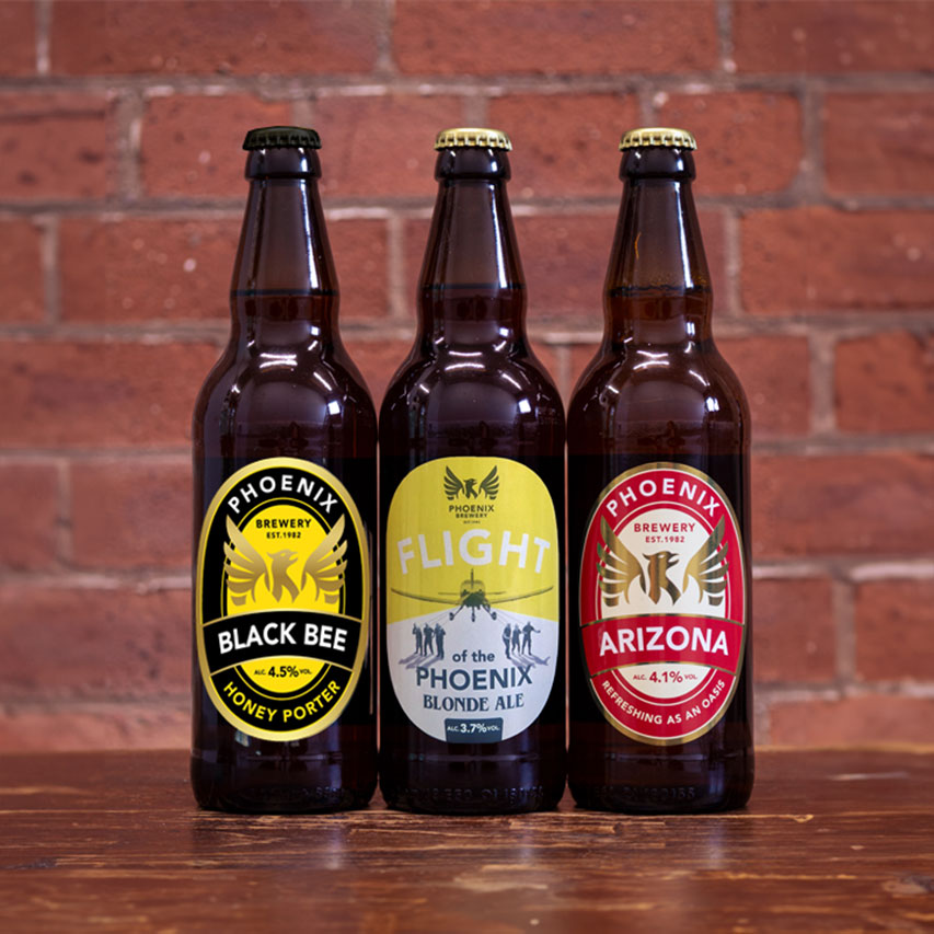 Three bottles of Phoenix Brewery beers lined up against a brick wall, featuring Black Bee, Flight Blonde Ale, and Arizona