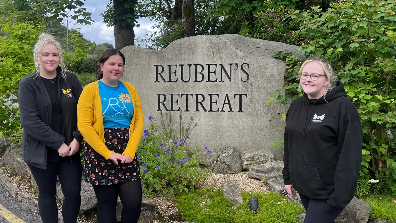 Lucy and Becca from Phoenix Brewery standing with Shannon in front of a large stone sign that reads 'Reuben's Retreat,' surrounded by greenery and flowers.