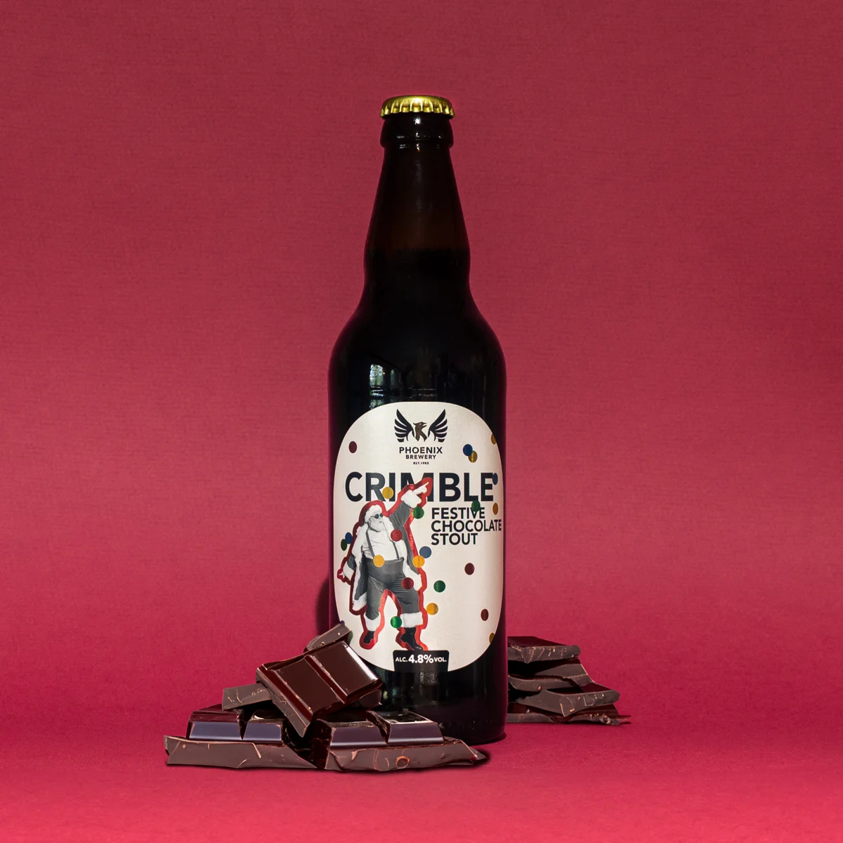 A bottle of Phoenix Brewery Crimble Festive Chocolate Stout surrounded by pieces of chocolate on a red background.