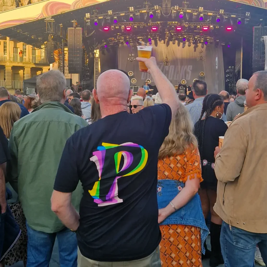 Person wearing a black P-logo t-shirt in a crowd at an outdoor concert, raising a beer