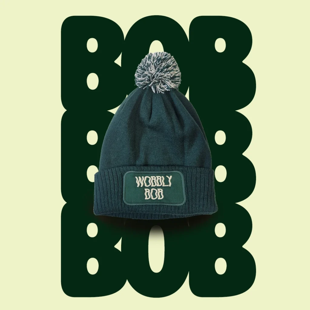 Dark green Wobbly Bob bobble hat with green-and-white pompom and embroidered patch logo on a cream background with dark green bubbly "BOB" text