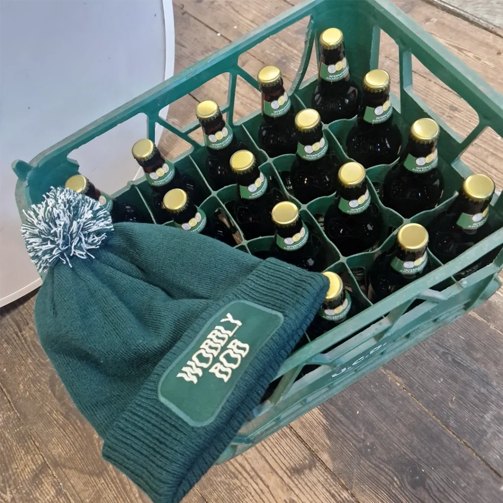 Green Wobbly Bob bobble hat with cream pompom resting against a crate of Wobbly Bob beer bottles on a wooden floor