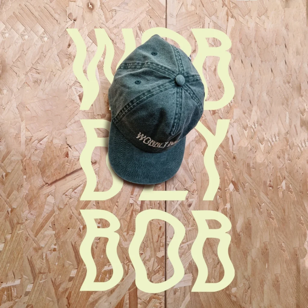 Washed dark green Wobbly Bob dad cap photographed from above on an OSB board background with large cream wavy "WOBBLY BOB" text