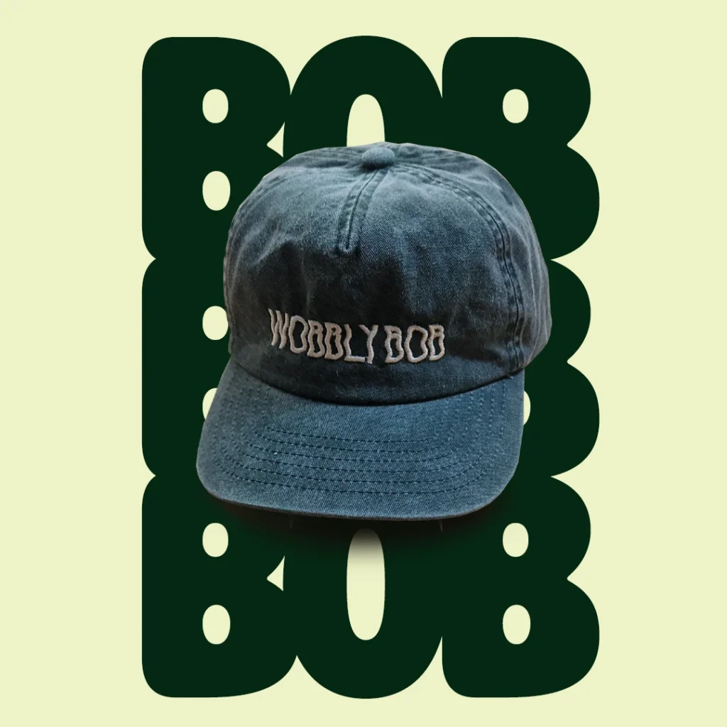 Washed dark green Wobbly Bob dad cap with cream embroidered logo on a cream background with large dark green bubbly "BOB" text