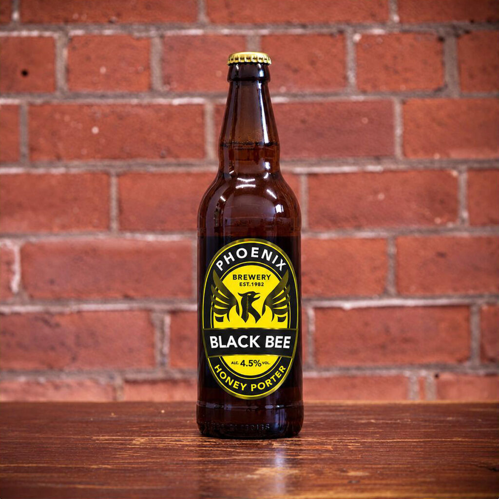 A bottle of Black Bee from Phoenix Brewery with a black and yellow label, placed on a wooden table with a brick wall in the background.