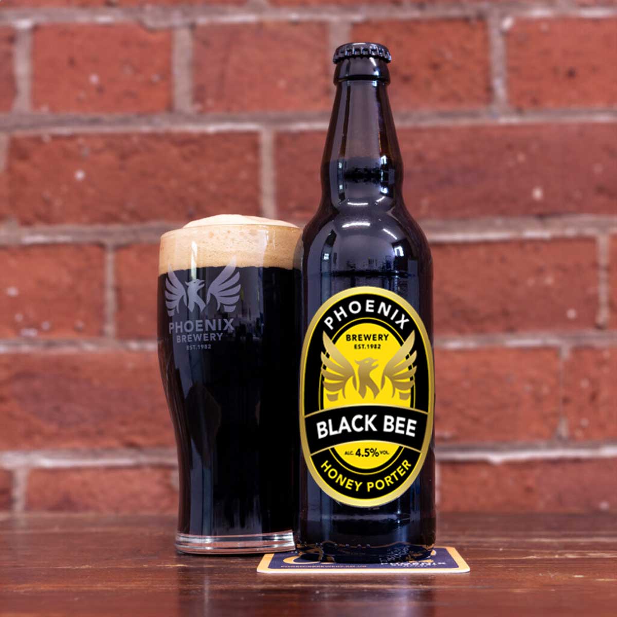 A bottle of Black Bee from Phoenix Brewery with a black and yellow label, next to a full pint glass, placed on a wooden table with a brick wall in the background.